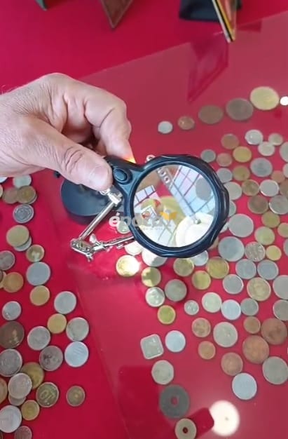 Mano con lupa sobre monedas de distintas épocas dispuestas sobre una superficie roja