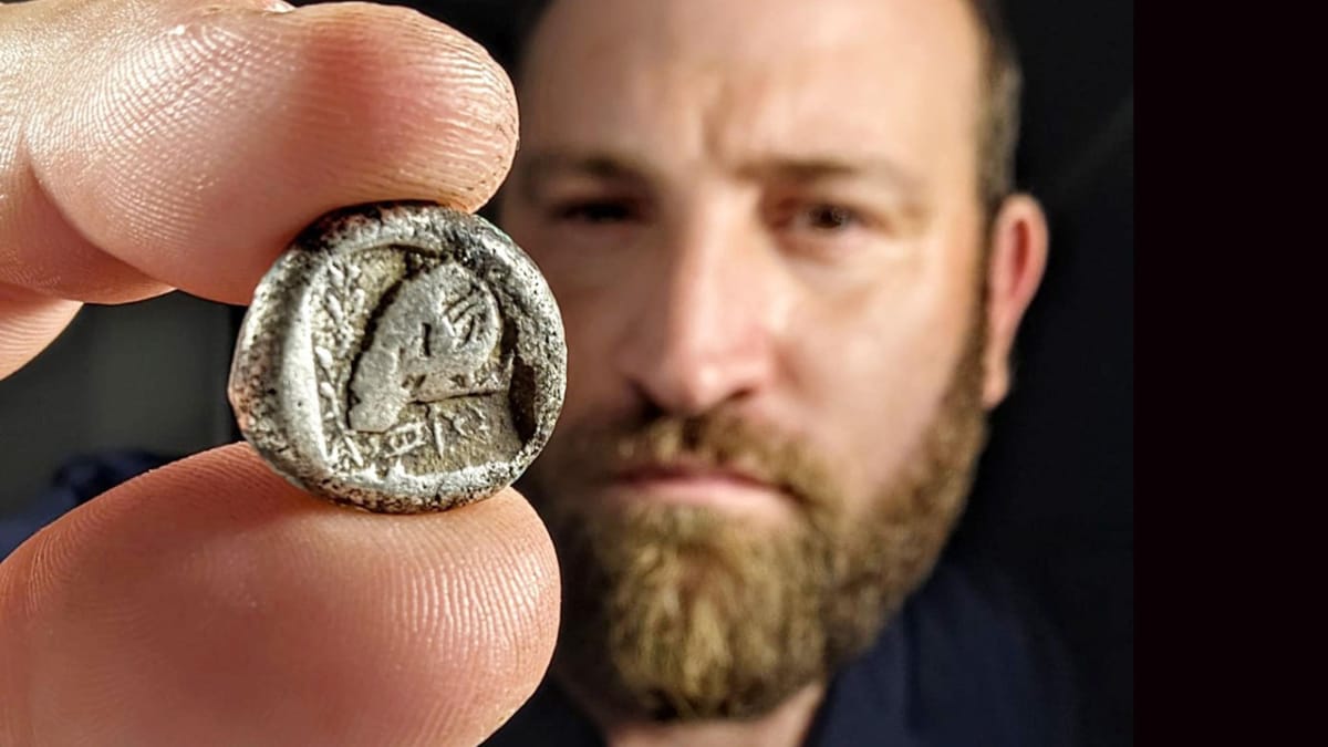 Moneda antigua de plata chipriota (siglos de Salamina) sostenida entre los dedos, con relieve de carnero; al fondo, un investigador observa la pieza