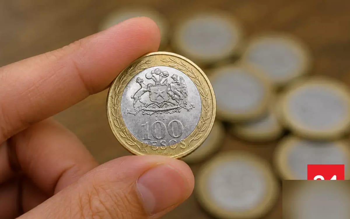 Mano sostiene una moneda bimetálica chilena de 100 pesos con el escudo nacional sobre otras monedas